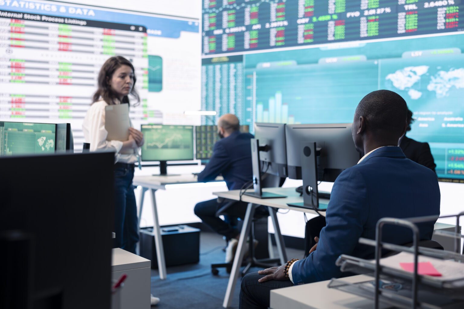 Professionals working in a financial office setting with graphs and financial market listings in the background.
